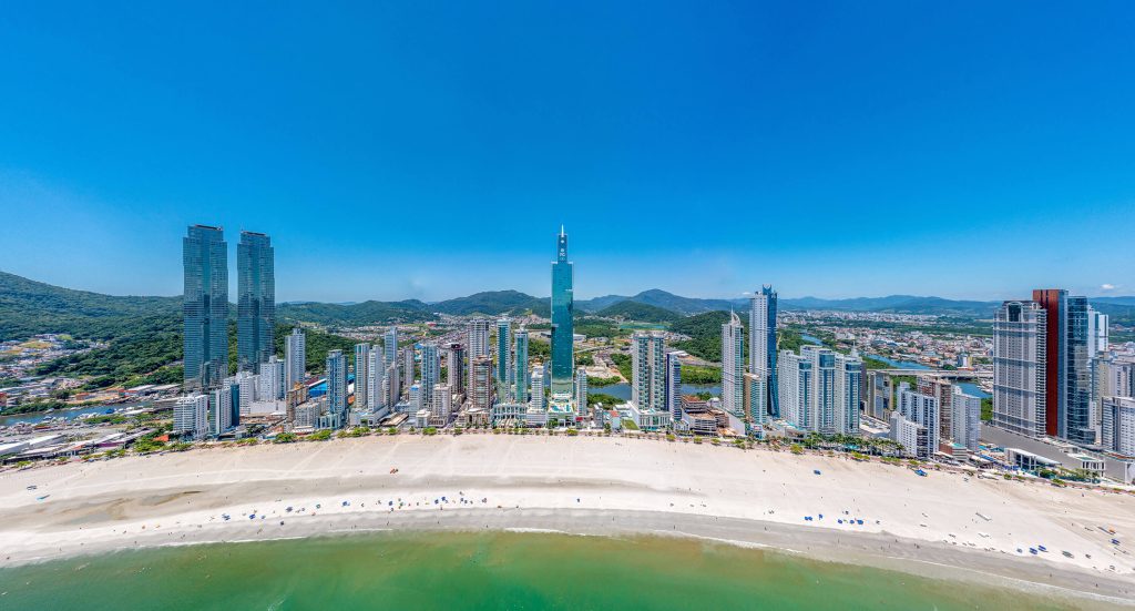 Vista ampla da orla de Balneário Camboriú com torres gêmeas e diversos arranha-céus voltados para o mar em um dia ensolarado.