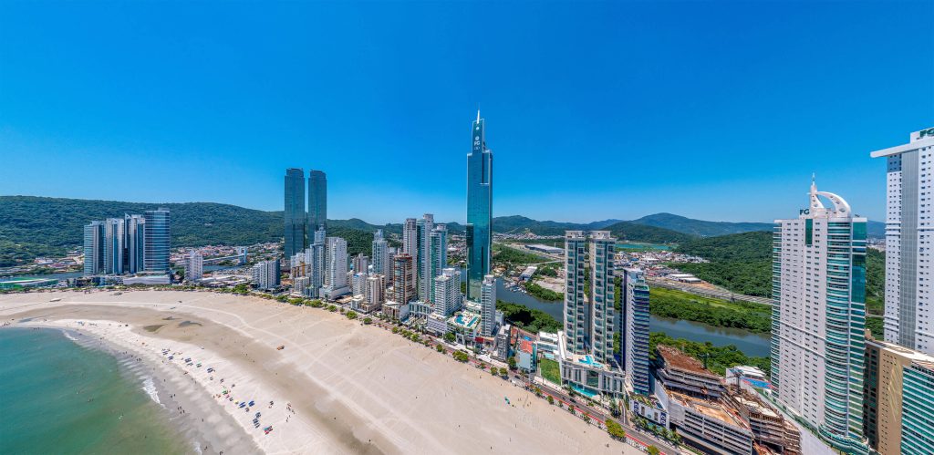 Vista ampla da orla de Balneário Camboriú com torres gêmeas e diversos arranha-céus voltados para o mar em um dia ensolarado.