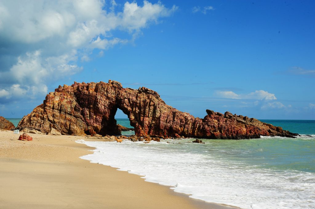 Pedra Furada em Jericoacoara (CE): arco de rocha avermelhada sobre faixa de areia clara e águas turquesa, com céu azul ao fundo