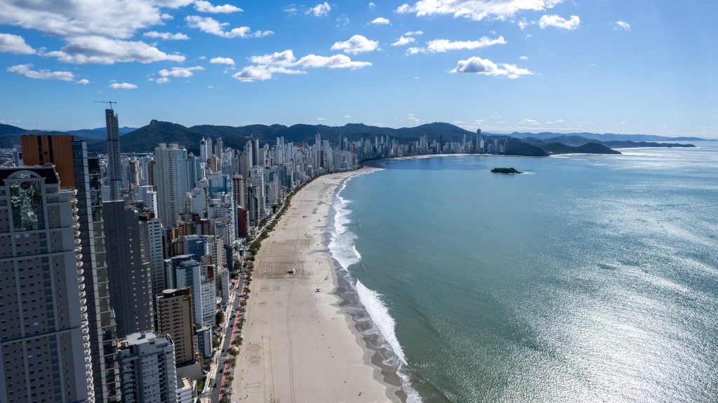Vista aérea de orla urbana com prédios altos alinhados à extensa faixa de areia e mar calmo sob céu parcialmente nublado.