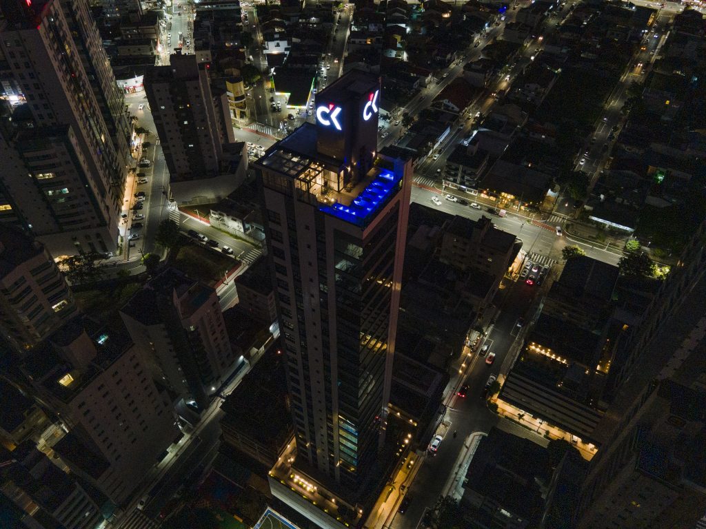 Vista aérea noturna de um arranha-céu com piscina e área de lounge iluminadas no topo, cercado por ruas e edifícios da cidade