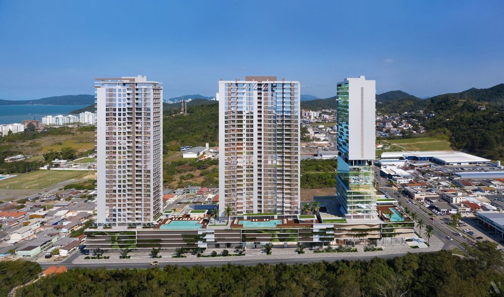 Visão frontal aérea do Home Clube Torres da Brava, com três torres modernas erguidas sobre um podium horizontal. No topo do podium, áreas de lazer com piscinas e espreguiçadeiras. Ao fundo, moradias baixas e a orla marítima da Praia Brava, com colinas cobertas de vegetação e um céu limpo de cor azul.
