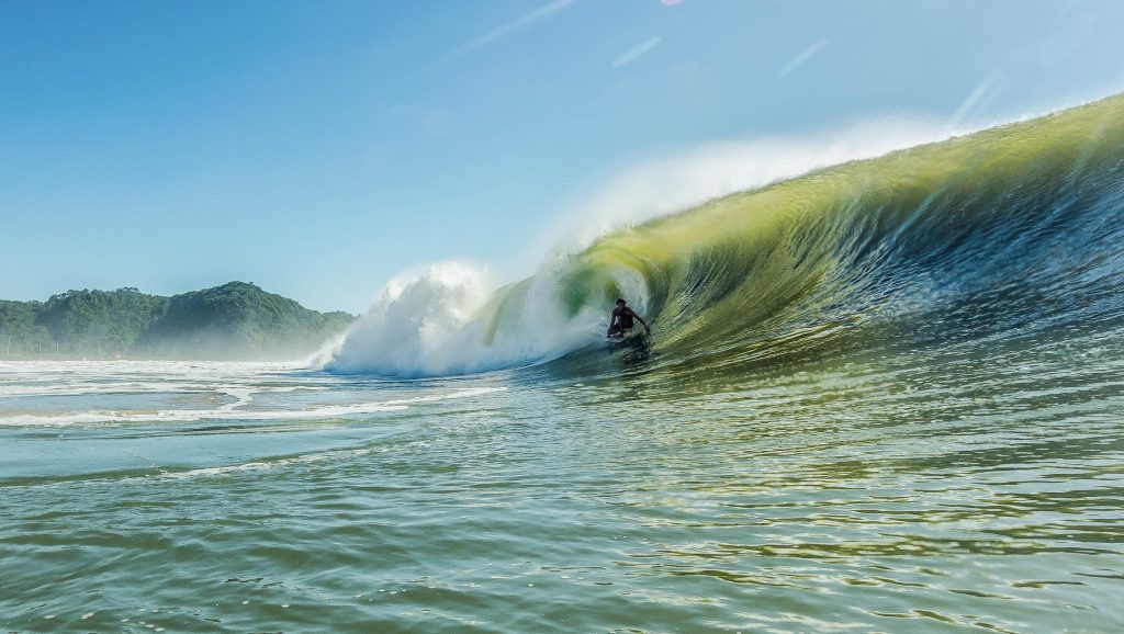 Surfista agachado dentro de uma onda tubular verde, com spray d’água iluminado pelo sol e céu azul ao fundo.