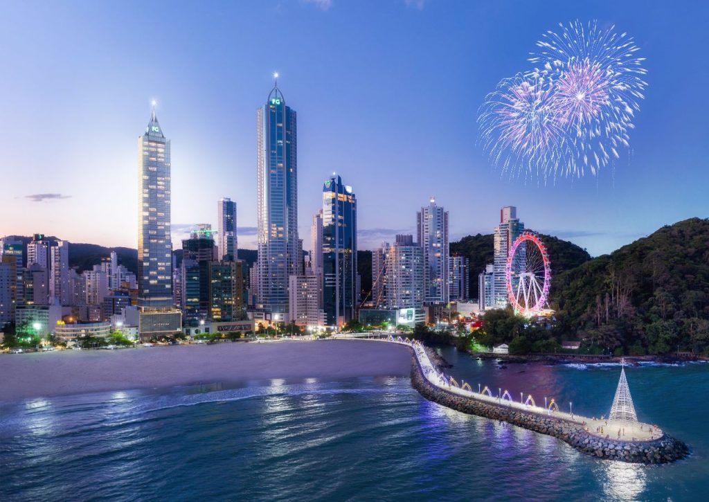 Paisagem urbana noturna de Balneário Camboriú apresentando arranha-céus iluminados, uma roda-gigante luminosa à beira-mar, fogos de artifício explodindo no céu e um píer de pedra iluminado estendendo-se pelo mar.