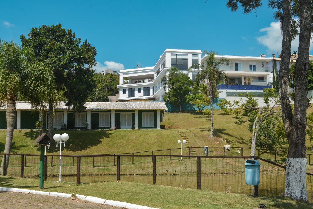 Residência de alto padrão em condomínio rural, com fachada branca, gramado, árvores e cerca junto a um lago pequeno.