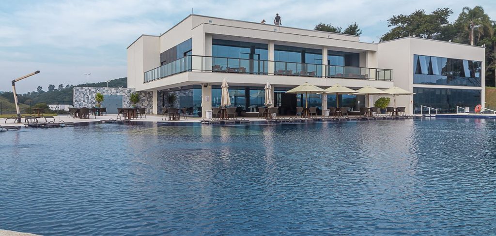 Clubhouse contemporâneo com fachada envidraçada e piscina de borda reta à frente, mesas com guarda-sóis na calçada.