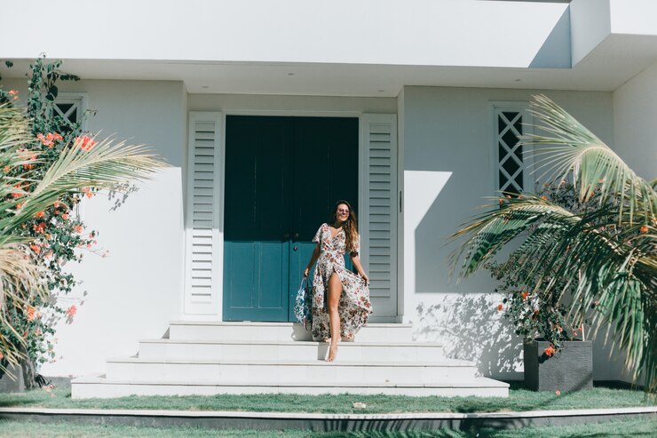 Mulher elegante em vestido floral descendo as escadas de entrada de uma casa de praia moderna, rodeada por folhagens tropicais