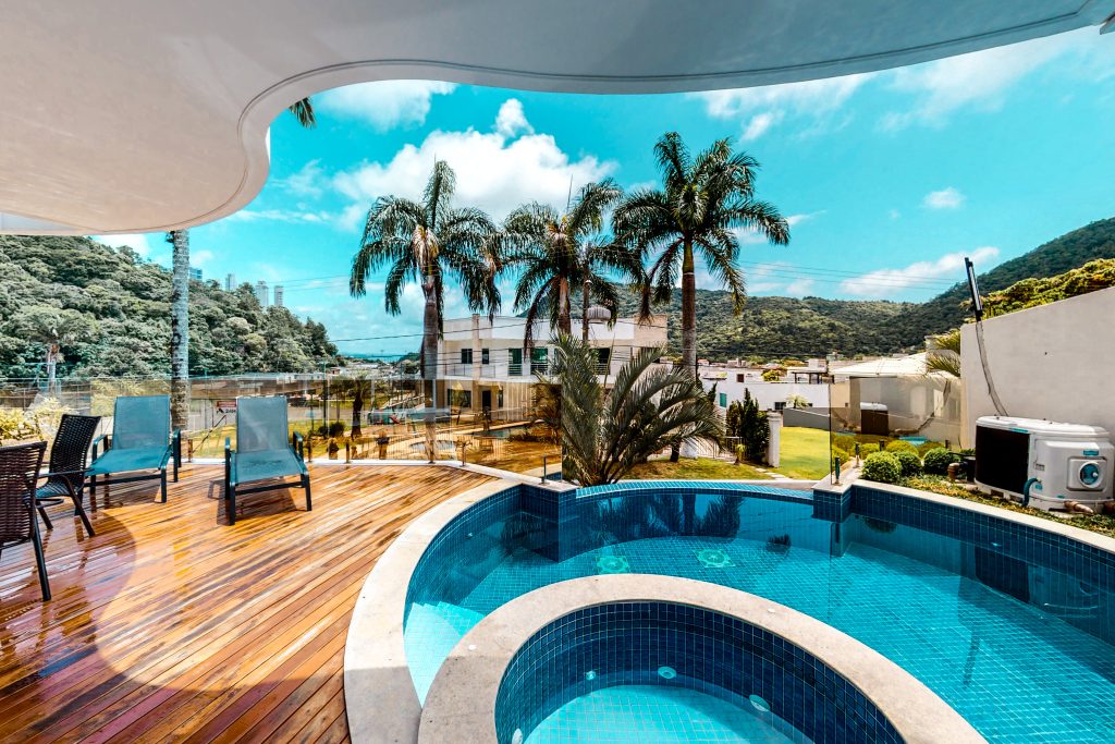 Deck de madeira com piscina em curva e espreguiçadeiras, vista parcial de jardins e encosta verde ao fundo.