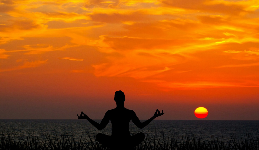 Silhueta de pessoa sentada em posição de lótus, braços abertos com as mãos em mudra, diante do mar calmo ao pôr-do-sol, com o céu pintado de tons laranja e dourado.