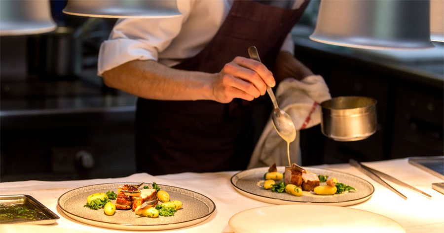 Chef em avental marrom, segurando uma concha de metal e com uma colher, finaliza dois pratos com porções de carne, batatas e folhas verdes sobre uma bancada iluminada.