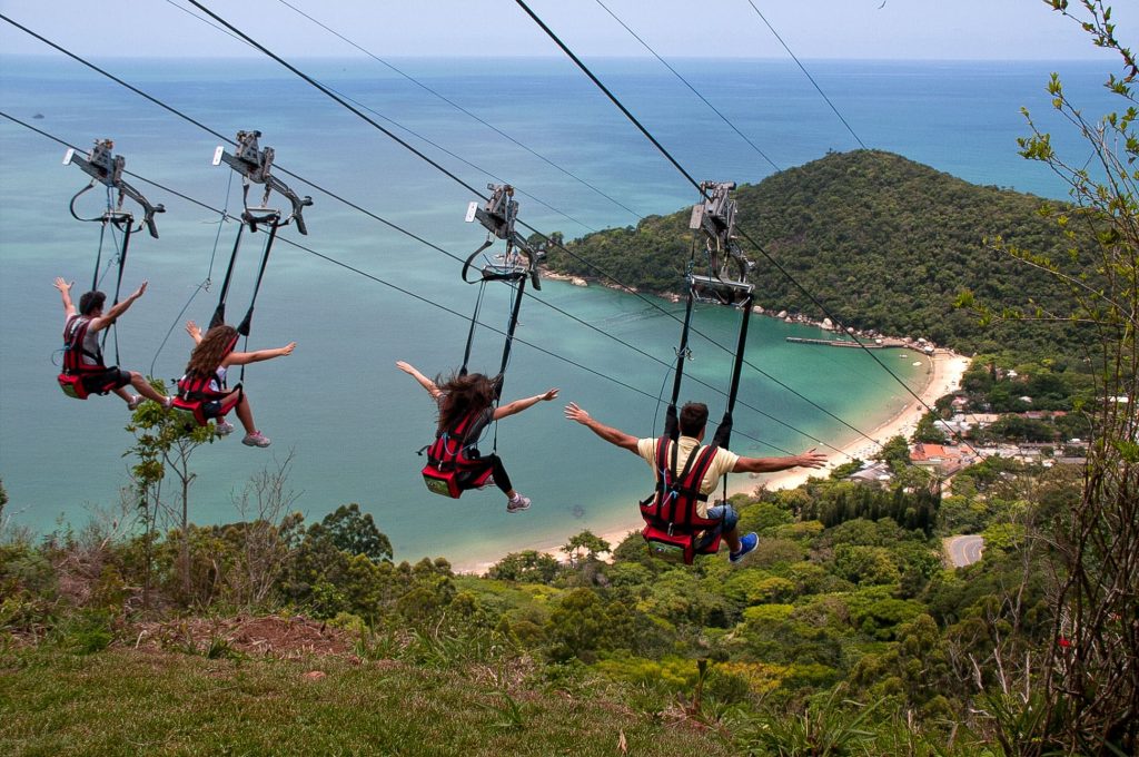 Turistas deslizando em cadeiras de tirolesa do Parque Unipraias com vista para o mar verde-turquesa e a Mata Atlântica ao fundo