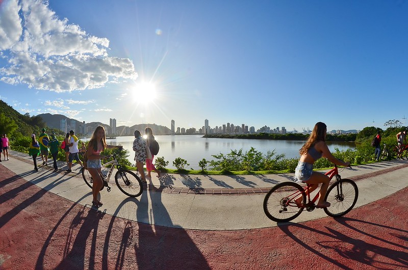 Ciclista em uma pista vermelha à beira de um rio, com pedestres e outros ciclistas ao lado, sol baixo no céu e o skyline de prédios ao fundo.