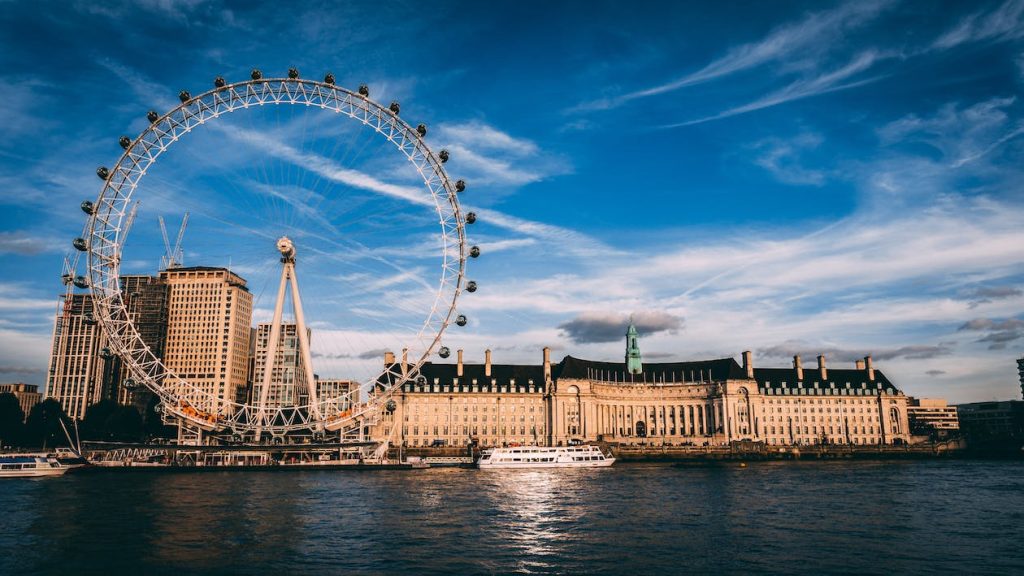 London Eye às margens do rio Tâmisa em frente ao edifício County Hall, com barcos navegando e céu parcialmente nublado ao entardecer.