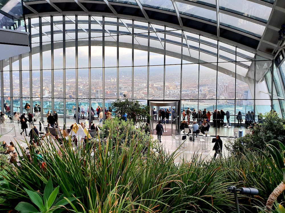 Deck de observação envidraçado em rooftop interno com plantas e visitantes apreciando vista panorâmica da cidade