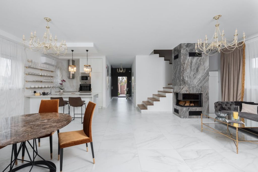 Ambiente integrado minimalista com piso de mármore branco, mesa de jantar redonda e cadeiras laranja, bancada de cozinha com banquetas, lustres em cristal, lareira em parede de mármore e escada de degraus flutuantes.