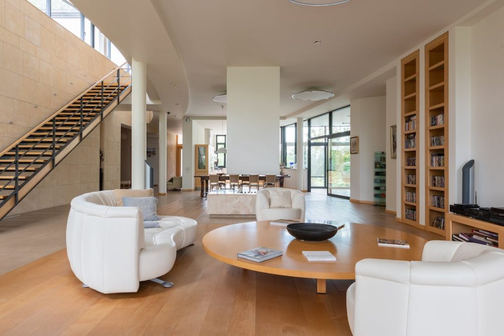 Sala duplex de alto padrão com pé-direito duplo, grandes janelas do chão ao teto com vista para um lago, sofá em “L” marrom, mesa de centro orgânica, plantas e escada para o mezanino.
