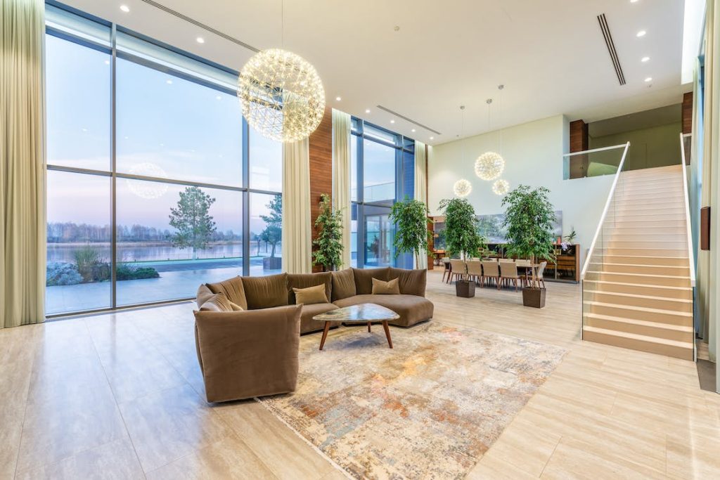 Sala duplex de alto padrão com pé-direito duplo, grandes janelas do chão ao teto com vista para um lago, sofá em “L” marrom, mesa de centro orgânica, plantas e escada para o mezanino.