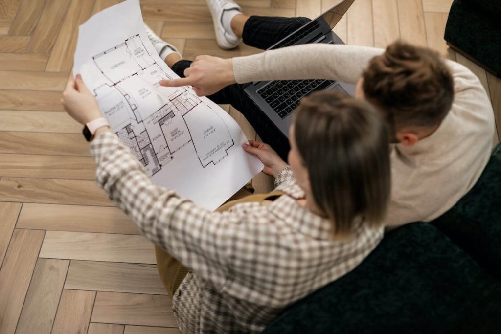 Casal sentado no chão analisando planta baixa de um apartamento com notebook ao lado