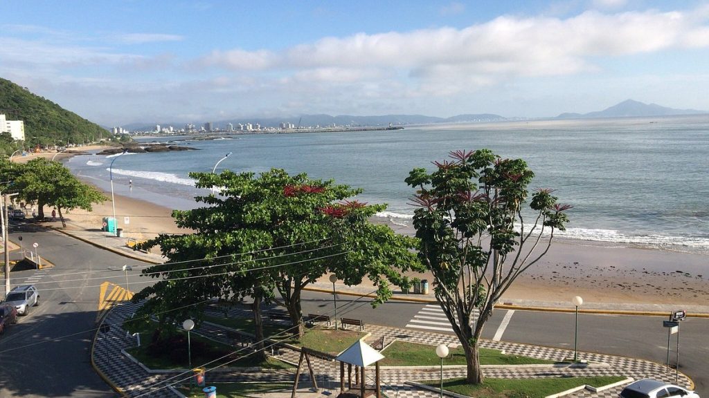 Vista da orla da Praia de Cabeçudas em Itajaí com calçadão em padrão xadrez, árvores e um pequeno playground em primeiro plano, avenida à beira-mar e o mar calmo com edifícios ao fundo.
