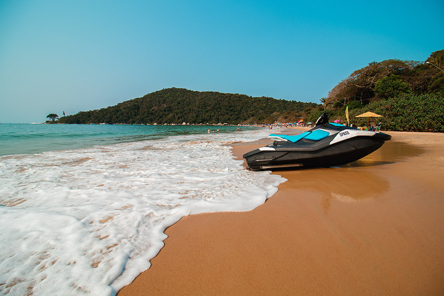 Jet ski estacionado na faixa de areia da Praia de Laranjeiras, com espuma suave das ondas na beira do mar e uma colina arborizada ao fundo.