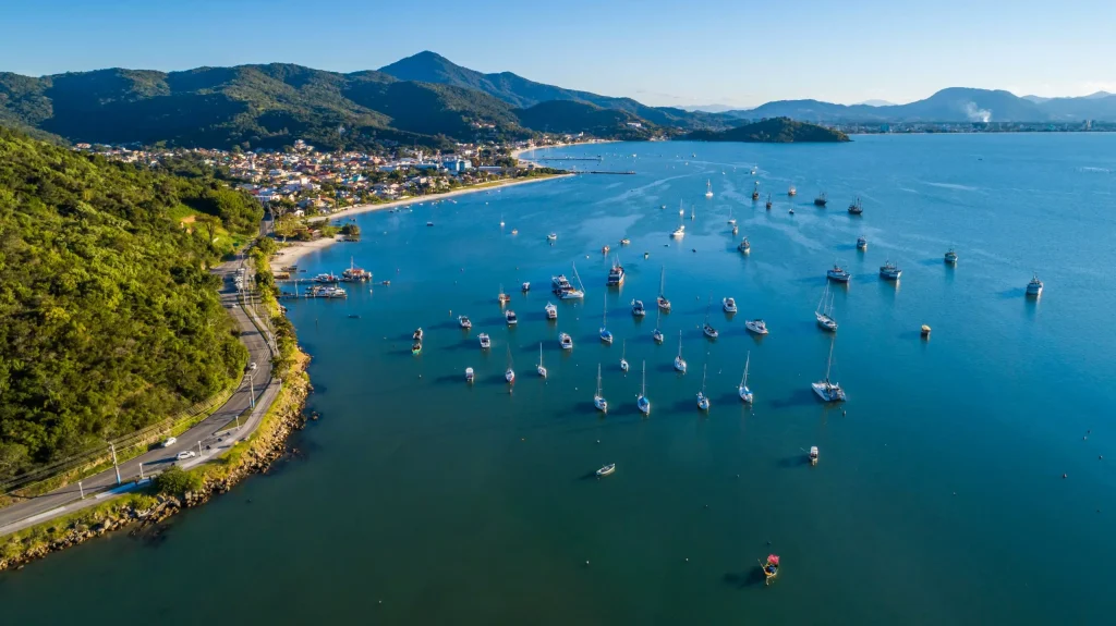 Vista aérea da Baía de Porto Belo: veleiros ancorados em águas calmas, estrada costeira contornando encosta verde e montanhas ao fundo.