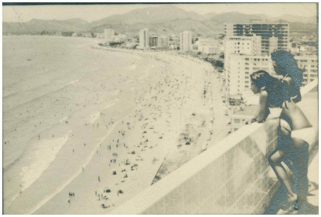 Fotografia antiga em preto e branco de duas jovens inclinadas sobre o parapeito de uma varanda, olhando para uma praia lotada abaixo e um horizonte urbano ao longe.
