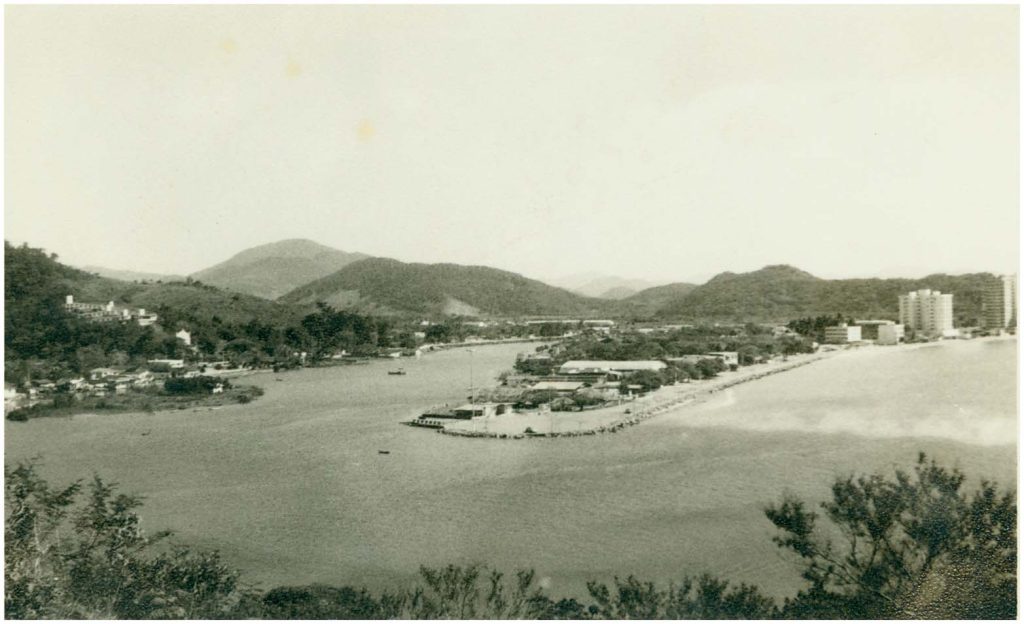 Foto em preto e branco histórica da Barra Sul em Balneário Camboriú, mostrando casas de veraneio à beira da água e um pequeno porto com colinas ao fundo.