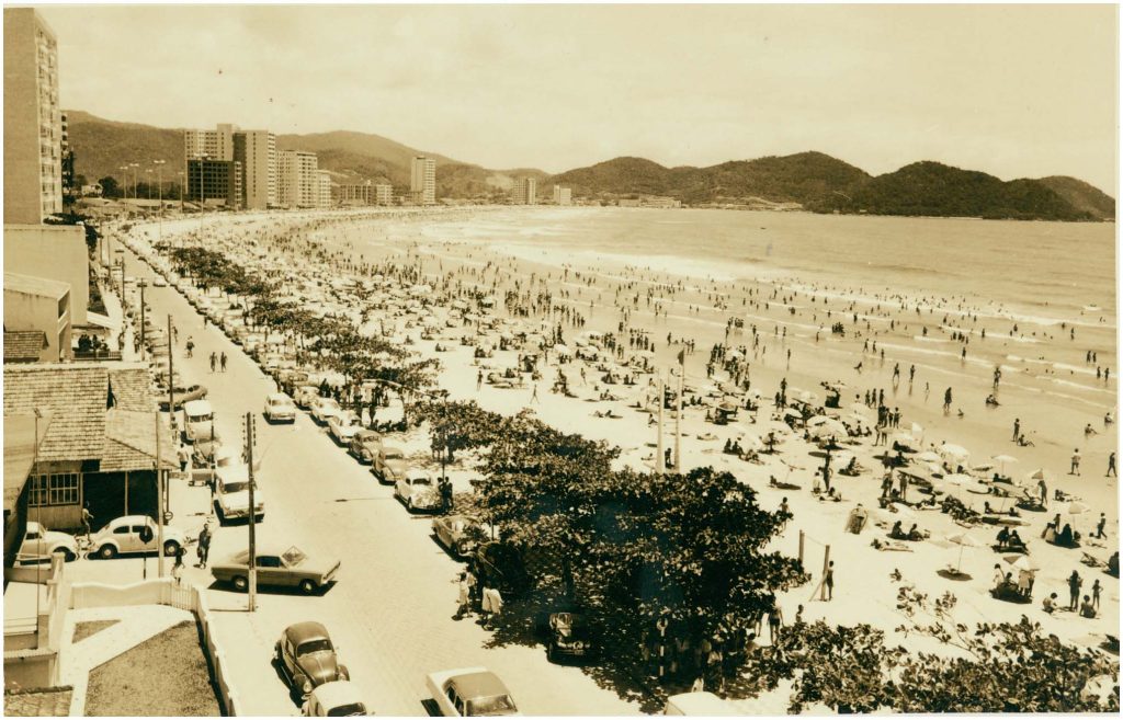 Imagem vintage sépia de uma avenida à beira-mar em Balneário Camboriú, com carros antigos estacionados e dezenas de banhistas espalhados pela faixa de areia.