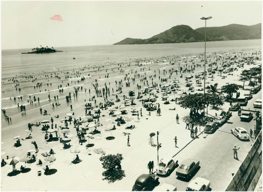 Foto em preto e branco de época antiga exibindo a praia de Balneário Camboriú lotada, com guarda-sóis, banhistas e carros clássicos estacionados ao longo do calçadão.