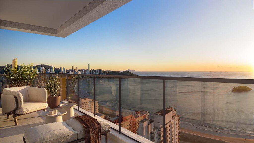 Varanda de apartamento de alto padrão com poltronas e chaise longue em tons neutros, guarda-corpo em vidro e vista panorâmica do litoral urbano e do oceano ao entardecer."