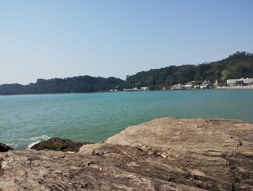 Formação rochosa plana em primeiro plano com águas calmas e esverdeadas da Praia de Cabeçudas; ao fundo, morro coberto de vegetação e edificações à beira da praia sob céu azul.