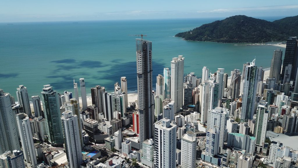 Vista aérea de orla urbana com numerosos edifícios altos brancos beirando o mar azul-turquesa e colina verde ao fundo, sob céu claro.