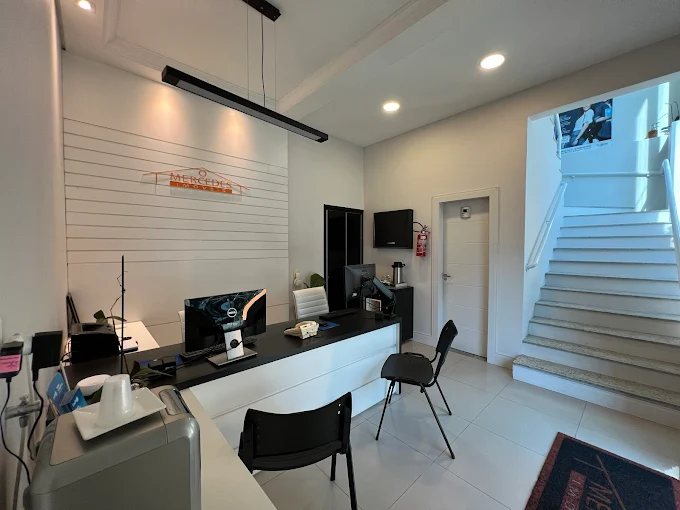 Recepção da Mercedes Imóveis em Itajaí (SC): balcão de atendimento branco com bancada preta, logo da imobiliária na parede, cadeiras de visita e escada ao fundo
