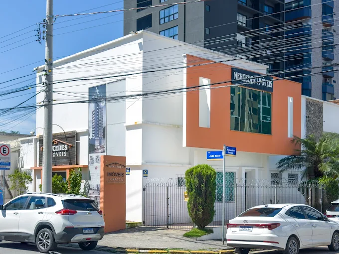Fachada da sede da Mercedes Imóveis em Itajaí (SC), prédio moderno branco com detalhes em laranja e letreiro da imobiliária.